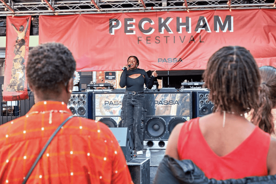 A woman on a stage with a microphone in her hand. "Peckham" is written in the background of the stage.